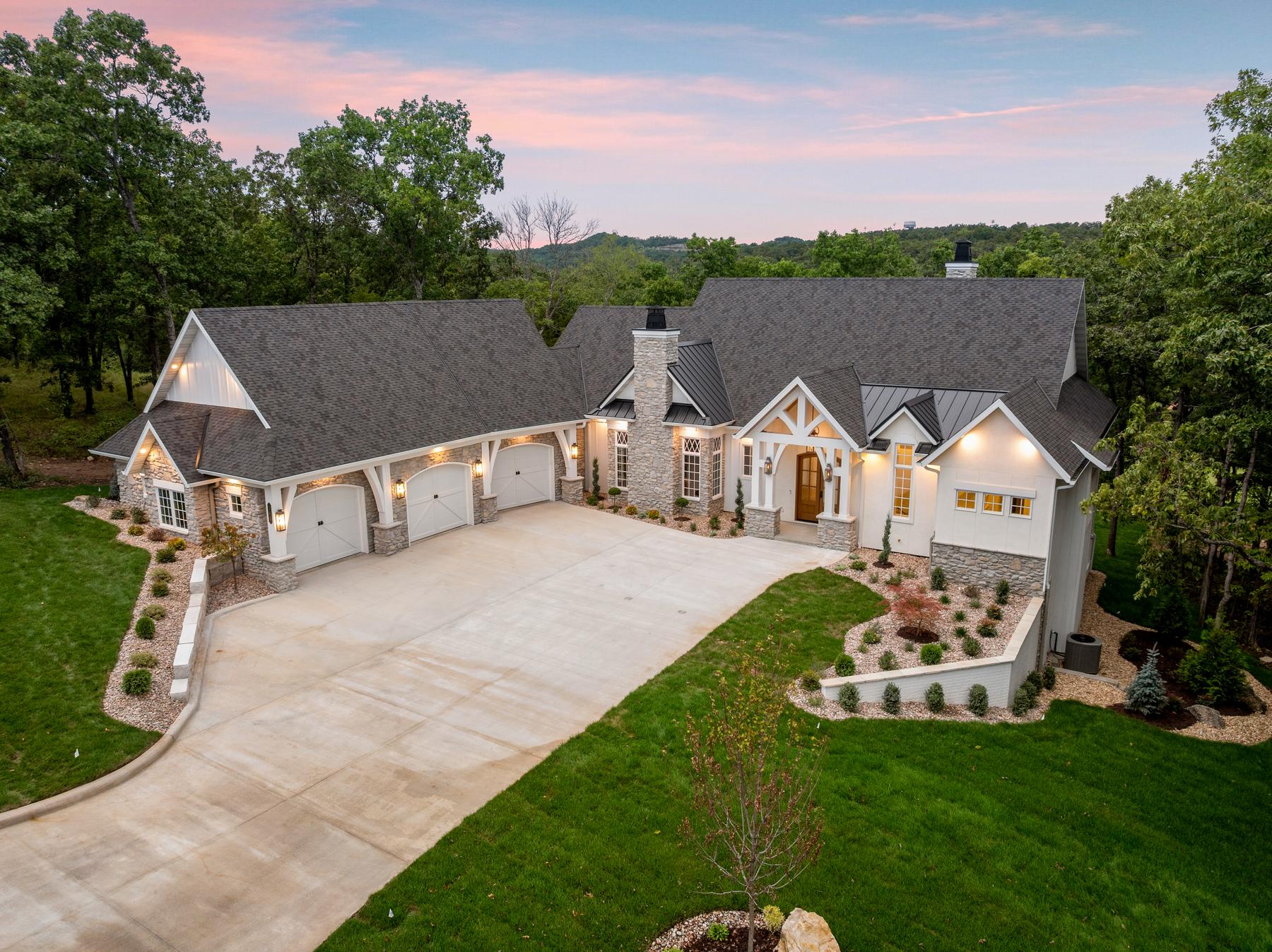Aerial view at dusk of a white craftsman home in Branson Hills with a stone chimney and a three-car garage