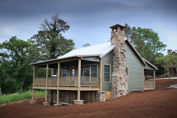 A green lakeside cabin with a stone chimney and a covered porch on two sides